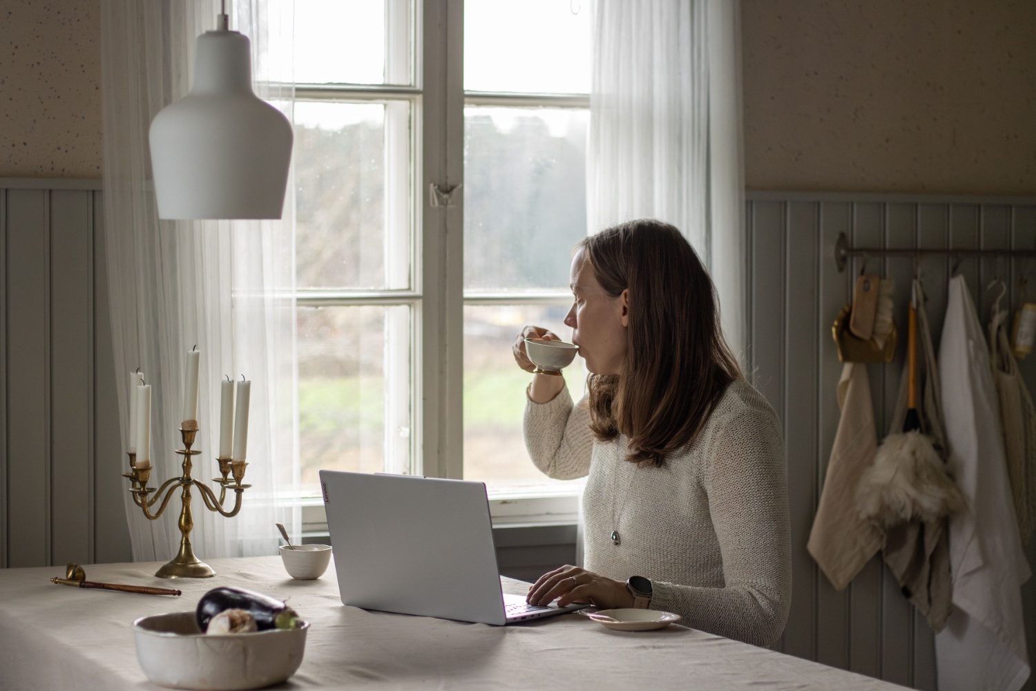 Nainen istuu pöydän ääressä vanhassa rakennuksessa juoden kahvia ja tehden töitä läppärillä.
